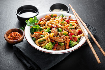 Udon stir fry noodles with pork meat and vegetables in a white plate on black stone background.
