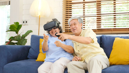 Senior asian couple playing virtual reality headset and using digital tablet in home living room with happiness emotion, retirement lifestyle and technology
