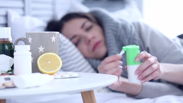 Beautiful sick ill woman female adult with scarf lying on bed with flu at grey bedroom with medicine, drugs and cup of tea with lemon, taking medicine against sore throat. Health issue concept.