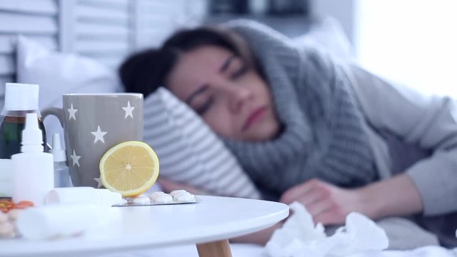 Beautiful sick ill woman female adult with scarf lying on bed with flu at grey bedroom with medicine, drugs and cup of tea with lemon, coughing and measuring temperature by electric thermometer.
