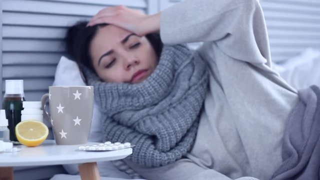 Beautiful sick ill woman female adult with scarf lying on bed with flu at grey bedroom with medicine, drugs and cup of tea with lemon, taking pill against cold. Health issue, flu and cold concept.