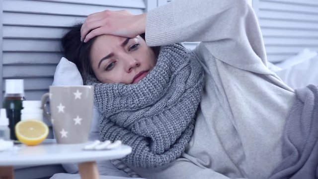 Beautiful sick ill woman female adult with scarf lying on bed with flu at grey bedroom with medicine, drugs and cup of tea with lemon, taking pill against cold. Health issue, flu and cold concept.