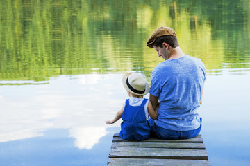 Dad and a tiny girl in dungarees relax at the river. Back view, copy space.