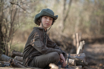 A boy in a coveralls and a hat sitting on a pile of sawn logs in the forest.