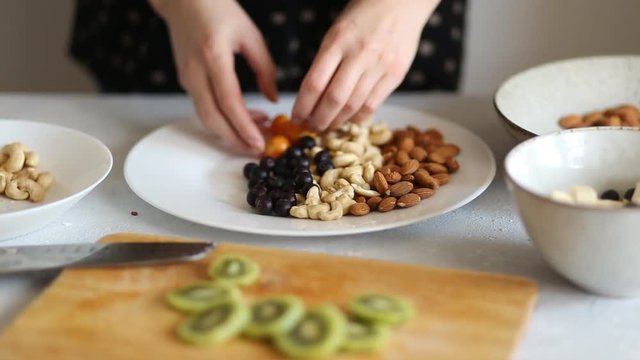 A woman prepares a delicious and healthy Breakfast, puts his hands cashew nuts, almonds, blueberries, kiwi and banana pieces in a large white dish, closeup