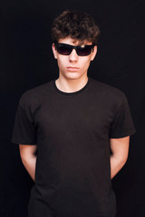 Fototapeta premium A boy of an adolescent age in a black shirt and in glasses from the sun on a black background. The boy has a serious face