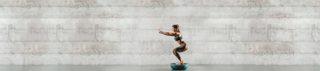 Side view of sporty woman doing exercises on bosu ball. In background gray wall, copy space. - Powered by Adobe