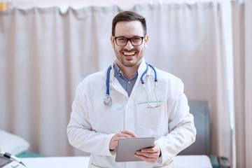 Cheerful Caucasian doctor in white uniform and with stethoscope around neck holding tablet an looking at camera while standing in hospital.