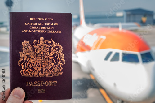 A red British passport held up against a background of a generic plane on a bright sunny day