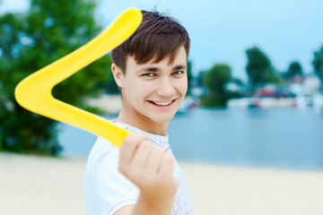 Happy young handsome active smiling man boy playing swings boomerang game on sand beech with blue river lake sky with clouds behind Concept of spring summer outdoor amusement activities entertainment - obrazy, fototapety, plakaty