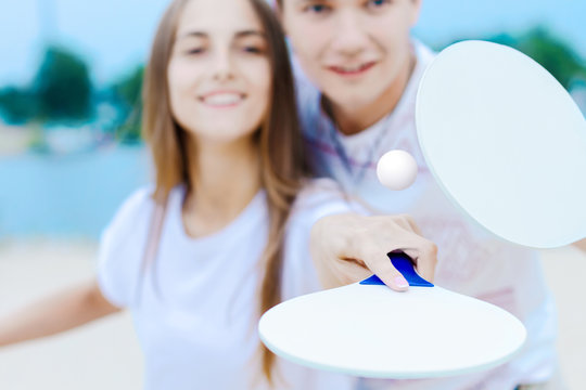 Young smiling man and woman in light clothes are kicking ball with white matkot rackets. Cheerful male and female are playing in tennis and having fun on beach on nature. Summer activities concept.
