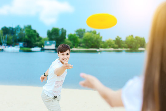 Happy active smiling romantic couple playing frisbee man throw disk game on sand beech with blue river lake sky with clouds behind Concept of spring summer outdoor amusement activities entertainment