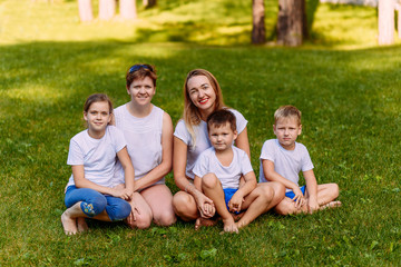 Fototapeta premium Two happy young mothers and three children in white t-shirts sit on a green lawn in the summer. Large Caucasian family on vacation