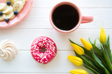 Black coffee and tasty doughnut, banana and blueberry snack on white rustic table, top view. Yellow flowers bouquet decoration. 
