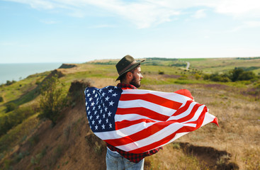 4th of July. Fourth of July. American with the national flag. American Flag. Independence Day. Patriotic holiday. The man is wearing a hat, a backpack, a shirt and jeans. Beautiful sunset light. 