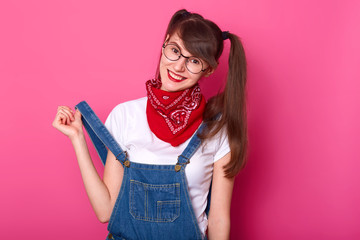 Playful smiling girl touches her denim overalls, being in high spirits. Cheerful long haired model poses isolated over bright pink background, wearing casual clothes and stylish accessories.