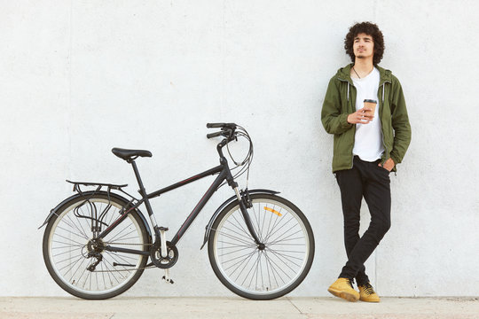 Indoor shot of attractive pencive man with crisp hair drinking hot baverage from paper cup, looks thoughtfully away, wears in fashionable clothes, stands near bicycle, isolated over white studio wall.