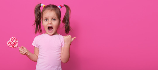 Emotional screaming little girl with pigtails shows direction with thumb, opens her mouth widely, opens eyes with terror, holding tasty sweet lollipop in other hand. Childhood and activities concept.