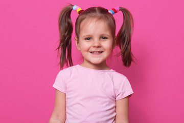 Close up portrait of cheerful sweet little girl with funny pigtails, smiling sincerely, looking directly at camera, standing straight, having colourful scrunchies. Copy space for advertisement.