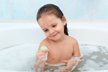 Happy baby taking bath alone, playing with foam bubbles and her new toy. Little child having fun in...
