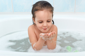 Shot of adorable baby girl with wet hair playing with soap foam in bath tub, has water procedure...