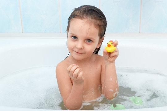 Close up portrait of happy charming little girl sitting in bath tub plays with yellow duck in bathroom. Baby bathing with toys in bath full of foam, showing toy to mother, looks seriously at camera