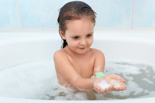 Funny baby girl playing with water and foam in big bath tub, has water procedures before going to bad, being in good mood, looking at her hands full of foam, wet baby having fun in bathroom.
