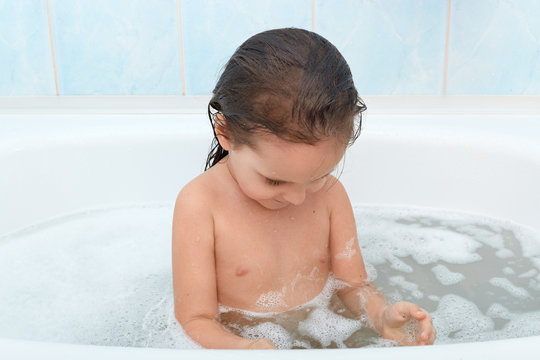 Image of bored little baby girl sits in bath tub, wants playing with duck in bathroom, waiting her mom with toys, baby bathing in bath full of foam before sleaping, lady looks down at foam in bath tub