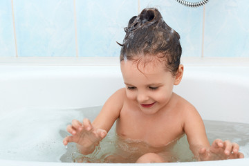 Cute baby having fun while taking bath, wet little girl sitting in bath tub, looking on water,...