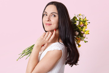 Attractive slender young lady holds flowers on her shoulder, touches chin with fingers, standing sidewards, looking another direction, wearing white summer dress, having bright red manicure.