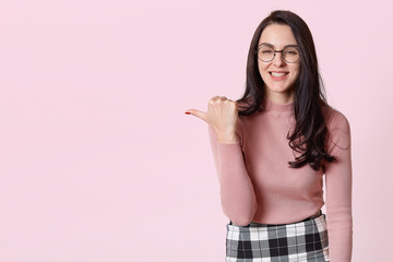 Positive smiling slender model standing over light pink background, showing direction with her thumb, laughing sincerely, wearing casual pink jumper, plaid skirt, fashionable round spectacles.