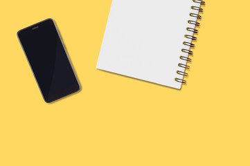 Black smartphone and blank paper notepad with spiral holder lies on yellow table in office or home. Top view. Business or education concept