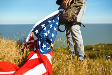 4th of July. Fourth of July. American with the national flag. American Flag. Independence Day. Patriotic holiday. The man is wearing a hat, a backpack, a shirt and jeans. Beautiful sunset light. 