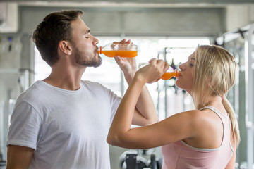 couple young fitness people drinking orange juice bottles in gym. sports man and woman exercises .
