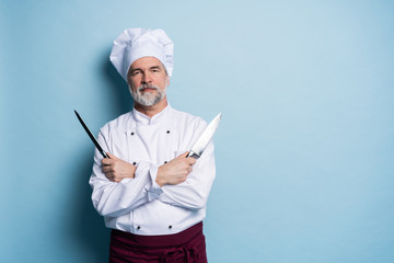 Portrait of a mature chef cook holding knifes isolated on a blue background.