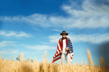 4th of July. Fourth of July. American with the national flag. American Flag. Independence Day. Patriotic holiday. The man is wearing a hat, a backpack, a shirt and jeans. Beautiful sunset light. 