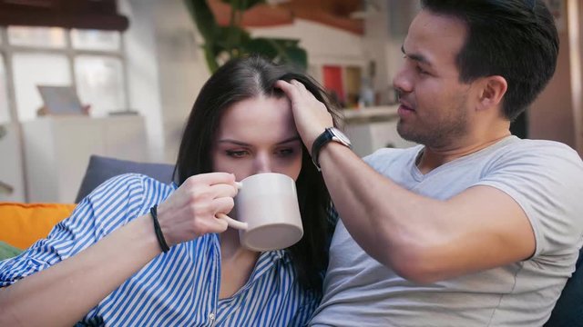 Cute Emotional Young Family Spend Pleasant Time Together, Man Taking Care Of His Wife, Gently Fixing Her Hair At Sofa In Nice Living Room, Girl Drink From Cup, Dressed In Grey T-shirt Light Blue Shirt