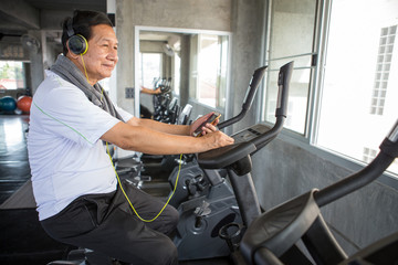 Senior older man  exercising on cycling machine listening music with headphones and phone relaxing...