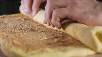 Pastry chef rolls the dough with cinnamon, sugar and butter into a roll on the table, selective focus. close-up.