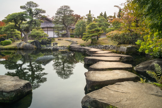 Perspective with large flat rocks forming a path over the pond and with a stone lantern and a Japanese building in the background, in autumn, at Koko-en Garden in Himeji, an Edo Style Japanese Garden 