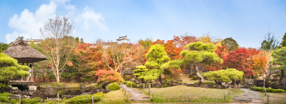 Chinese style garden panorama in autumn at Koko-en Japanese Gardens with small stone bridges over the creek and with the rooftop of Himeji Castle just peeking over the colorful tree line.