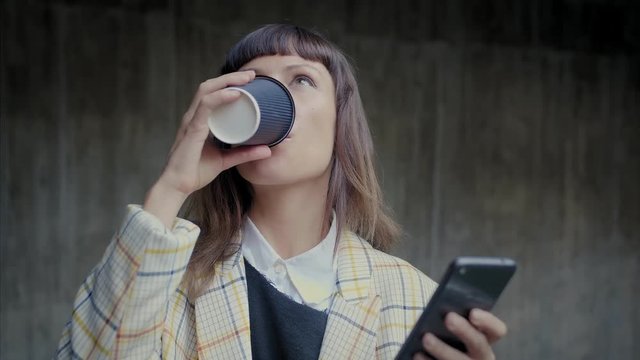 Portrait of Caucasian emotional business female enjoying coffee in take away cup and mobile phone device in both hands, dressed suit and shirt, using social media application, reading online feed