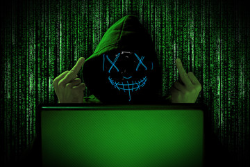 hacker with blue glowing mask behind notebook laptop in front of green source binary code...