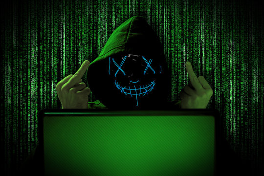 hacker with blue glowing mask behind notebook laptop in front of green source binary code background internet cyber hack attack concept