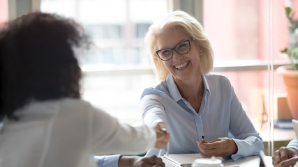 Happy mature caucasian leader handshake african employee at group meeting