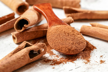 Wooden scoop with ground cinnamon powder, whole bark sticks in background on white baord