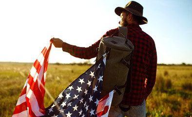 Naklejka premium 4th of July. Fourth of July. American with the national flag. American Flag. Independence Day. Patriotic holiday. The man is wearing a hat, a backpack, a shirt and jeans. Beautiful sunset light. 