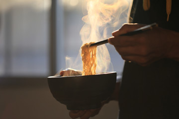 Man hand holding chopsticks of instant noodles in wood bowl with smoke rising in the home, Sodium...