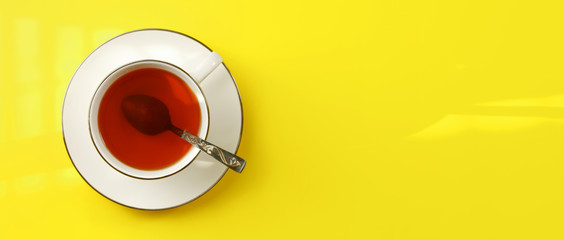 Flat lay photo - white porcelain cup with spoon filled hot amber tea, on yellow board, wide banner...
