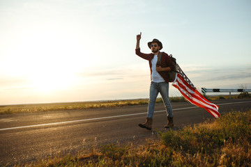Fototapeta na wymiar 4th of July. Fourth of July. American with the national flag. American Flag. Independence Day. Patriotic holiday. The man is wearing a hat, a backpack, a shirt and jeans. Beautiful sunset light. 
